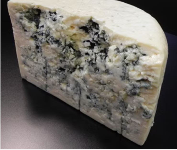 A half-cut wheel of Big Rock Blue cheese, showcasing its ivory-colored interior with blue veins, is set against a dark grey background