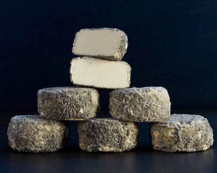 Stacks of Brightwell Ash cheese discs, with a few half-cut pieces on top revealing their pale white interior and ash-coated rind, set against a black background