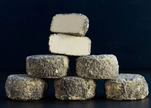 Stacks of Brightwell Ash cheese discs, with a few half-cut pieces on top revealing their pale white interior and ash-coated rind, set against a black background