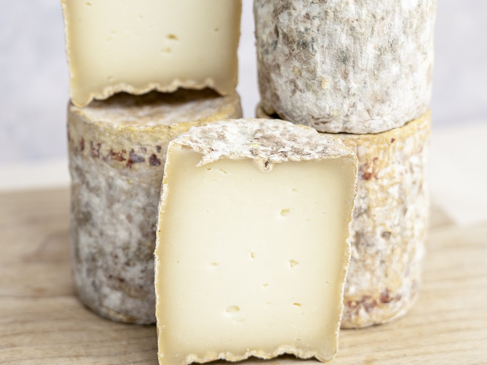Barrel-shaped Barriquet cheeses are stacked on a wooden surface, with some partially cut to reveal their smooth interior and washed rind