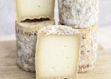 Barrel-shaped Barriquet cheeses are stacked on a wooden surface, with some partially cut to reveal their smooth interior and washed rind