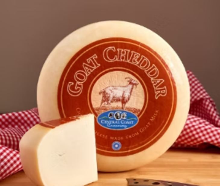 A disc of Goat Cheddar, wrapped in a sheet with a label on top, sits on a wooden board alongside a slice of the cheese, a cutting knife, and a red-checked cloth.
