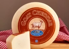 A disc of Goat Cheddar, wrapped in a sheet with a label on top, sits on a wooden board alongside a slice of the cheese, a cutting knife, and a red-checked cloth.