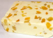 A piece of White Stilton infused with Mango & Ginger cheese, showcasing mango pieces, rests on a grey background.