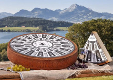 A wheel of Le Gruyère AOP Cheese 168开奖官方开奖结果查询, accompanied by a triangular slice with a label on top, placed on a wooden board with a scenic backdrop of mountains and a river