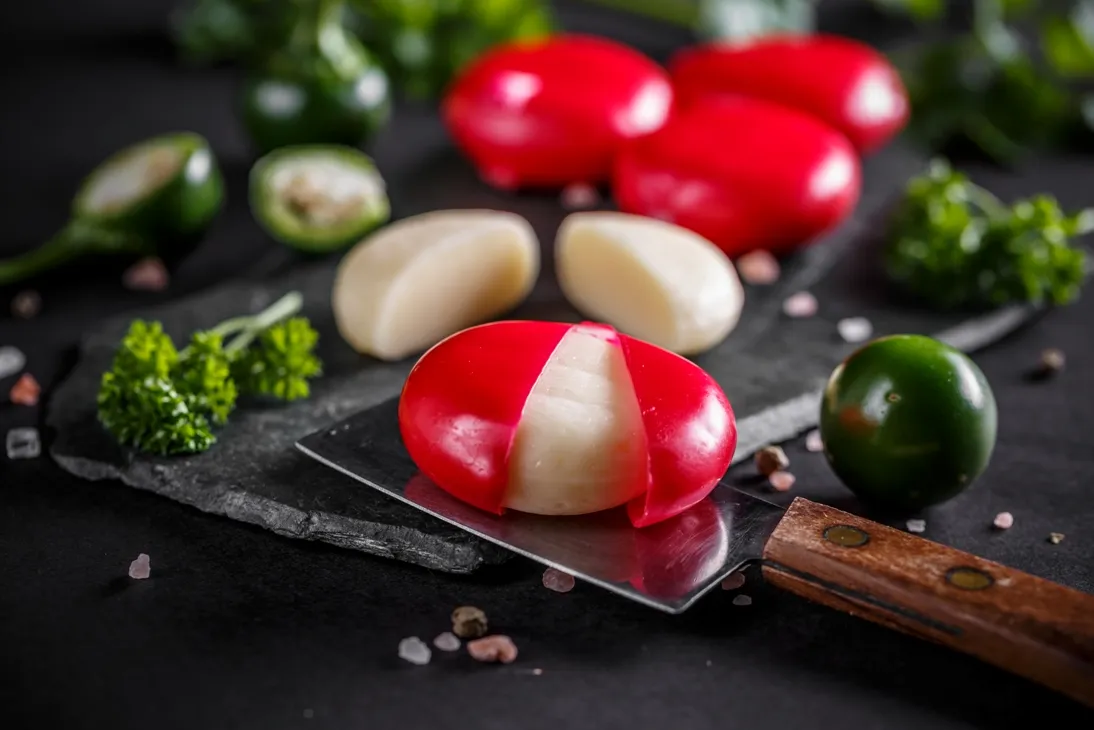 Small rounds of Babybel cheese with red wax, arranged on black marble alongside fresh parsley, green olives, and a cutting knife