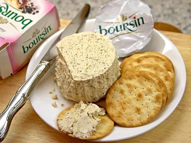 Boursin cheese spread on a cracker, with a large portion of Boursin cheese, on a white plate next to a box of the same cheese and a knife