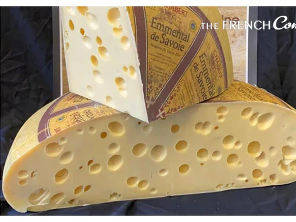 An Emmental cheese wheel, cut in half to reveal its large holes, with a slice of Emmental cheese placed on top of the cut portion