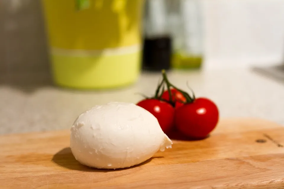 A fresh ball of Fior Di Latte cheese sits on a wooden cutting board, with a small bunch of ripe red tomatoes and kitchen items in the background creating a cozy atmosphere.