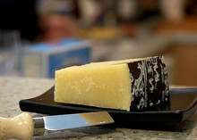 A black plate showcases a piece of Pecorino Romano Cheese 168开奖官方开奖结果查询 幸运8官网开奖官方结果 直播历史记录查询 with its protective covering on one side, revealing its granular texture, accompanied by a knife